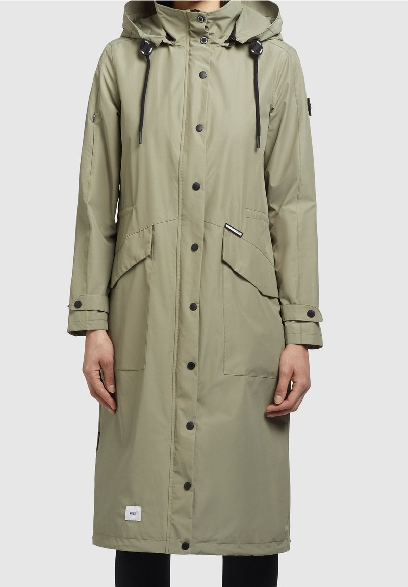 Imperméable long vert olive, fabriqué en tissu lisse et résistant à l'eau. Comprend une capuche, des boutons-pression, deux grandes poches avant et des poignets ajustables.
