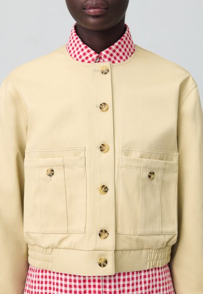 Veste cropped beige clair avec deux poches avant, boutons dorés et col écossais rouge et blanc. Tissu lisse avec une ceinture élastique.