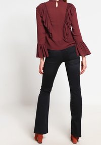 Femme portant un blouse bordeaux à volants avec des manches évasées, un jean noir évasé, et des bottes à talons orange brûlé, se tenant dos à la caméra.