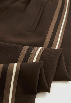 Pantalones deportivos marrones con rayas horizontales en tonos de beige, crema y marrón oscuro. Están hechos de un tejido suave y texturizado con bolsillos.