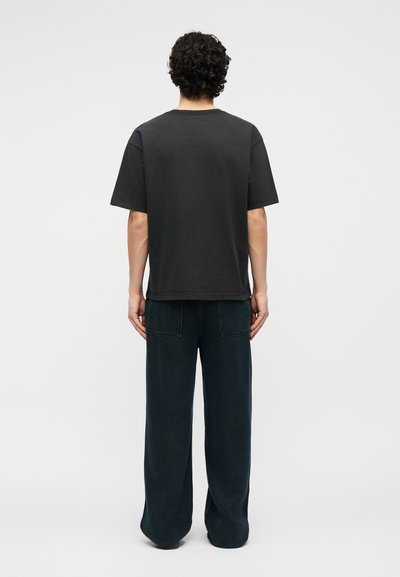 Άνδρας με σγουρά μαλλιά, που φοράει μαύρο κοντομάνικο πουκάμισο και άνετο μαύρο παντελόνι, στέκεται με τα χέρια δίπλα στο σώμα, γυρισμένος πλάτη σε απλό φόντο.