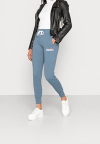 Blue joggers with white drawstring, elastic cuffs, and logo on the thigh, paired with a white turtleneck and black leather jacket. White sneakers.