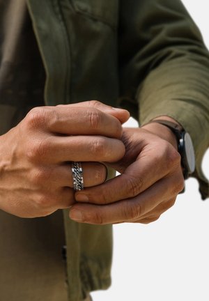 Silver rings on a hand, one with a chain pattern and the other smooth. The hand's skin tone is medium. An olive green jacket is partially visible.