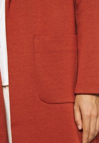 Rote, strukturierte Jacke mit einer seitlichen Tasche und entspanntem Schnitt. Der Stoff wirkt weich und leicht. Ein Arm ist teilweise sichtbar, die Hand ruht.