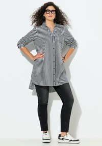 Camicia lunga a righe bianche e nere con chiusura a bottoni, orlo arrotondato e taschino sul petto, abbinata a jeans skinny neri e sneakers bianche e nere.