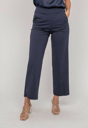 Femme portant un pantalon large bleu marine avec poches latérales et des sandales à talons hauts dorées à brides, debout devant un fond uni.