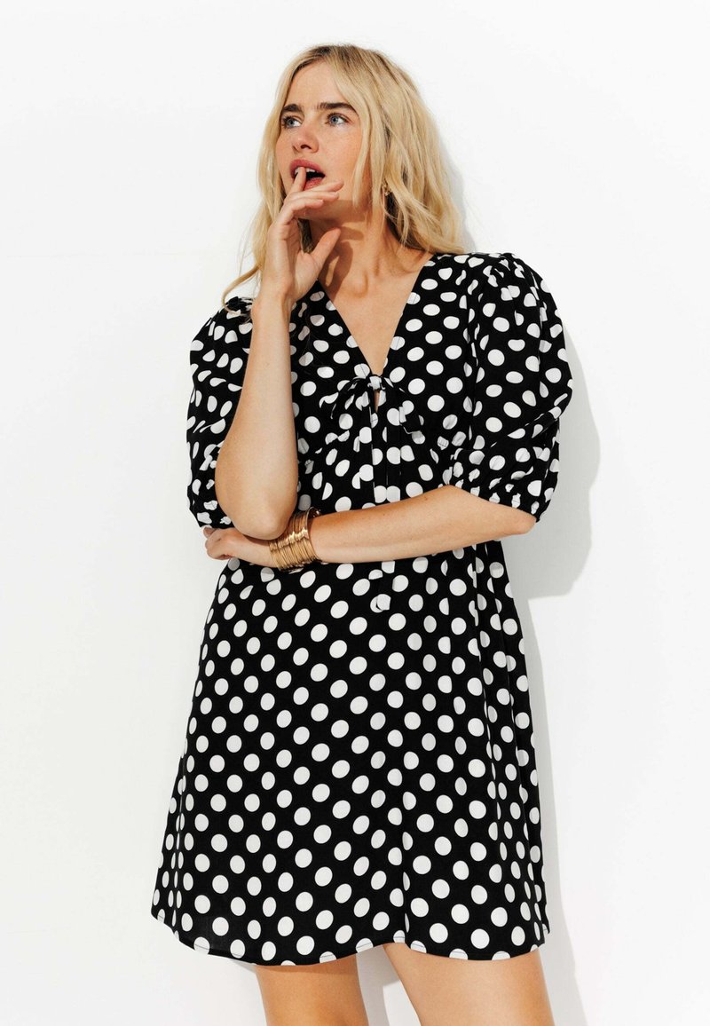 Robe noire à pois blancs, décolleté en V, manches bouffantes et détail noué à l'avant. Tissu doux avec une silhouette fluide.