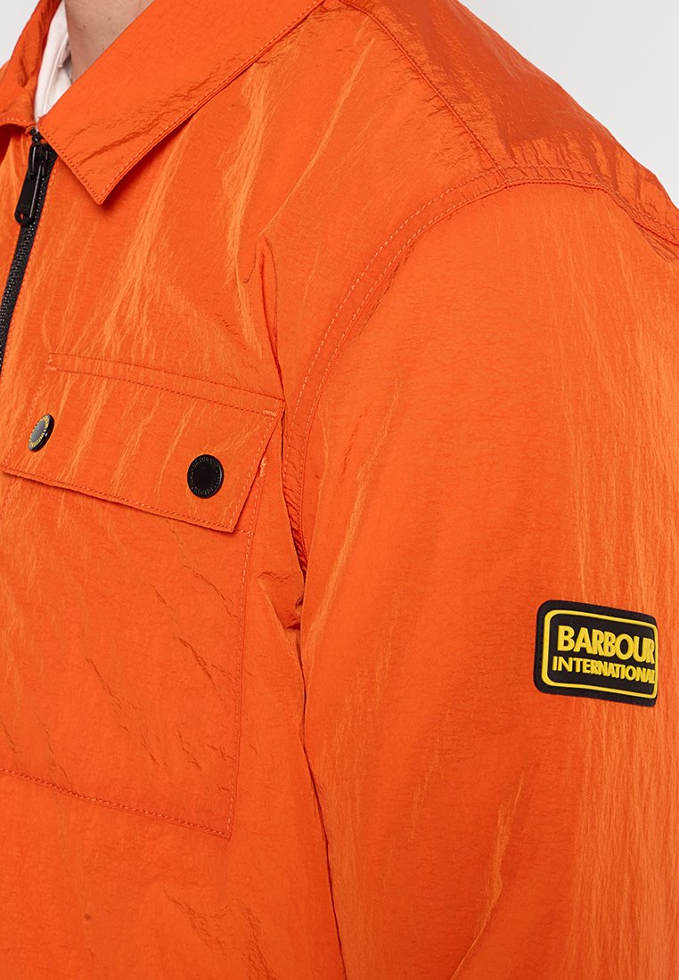 Orangefarbene Jacke aus leichtem Material, mit einer geknöpften Brusttasche, schwarzem Zubehör und einem sichtbaren Markenlogo am Ärmel.