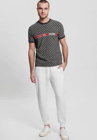 Zwart t-shirt met patroon en rood-wit "GUESS" logo, gecombineerd met witte joggers en witte sneakers. Casual, sportieve uitstraling.