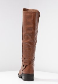 Botte en cuir marron, haute, avec un petit talon empilé, une sangle décorative et un détail en métal, vue de derrière sur une surface blanche.