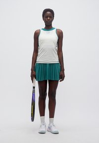 Γυναίκα τενίστρια στέκεται κρατώντας ρακέτα στο δεξί χέρι, φορώντας λευκό αμάνικο τοπ, τυρκουάζ πλισέ φούστα, λευκές κάλτσες και αθλητικά παπούτσια.