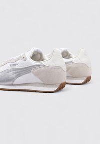 Zapatillas blancas con un detalle lateral texturizado en plata, talón de ante y suela de goma. Cuenta con un cuello acolchado y cordones delgados.