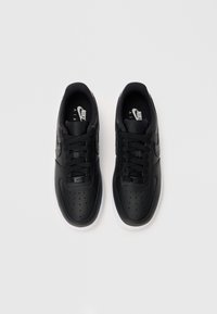 Par crnih Nike Air Force 1 tenisica s vezicama, bijelim potplatima i perforiranim prednjim dijelovima, prikazanih odozgo na svijetloj pozadini.