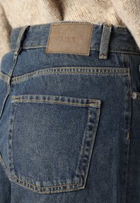 Denim kavbojke z usnjeno etiketo z napisom "JUXX," ki imajo tradicionalno zadnjo žepek in modro tkanino z teksturiranim zaključkom.