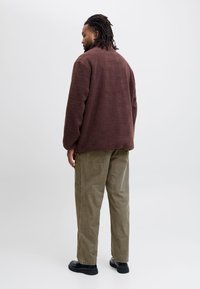 Bruine fleece jas met een ontspannen pasvorm, gecombineerd met olijfgroene corduroy broek en zwarte schoenen. Op een egale witte achtergrond.