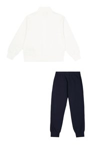 Weiße Zip-Jacke mit hohem Kragen und gerippten Bündchen, kombiniert mit navyblauen Hose mit elastischem Bund und umgekrempelten Säumen. Der Stoff wirkt glatt.