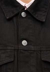 Μαύρο τζιν μπουφάν με κλασικό γιακά, μπροστινή τσέπη και μεταλλικά κουμπώματα. Το ύφασμα έχει υφή με αντίθετη ραφή.