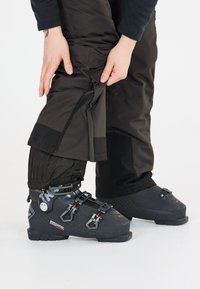 Svarta skidboots med metallspännen och remmar, tillsammans med mörka byxor som har dragkedjor och en strukturerad tyg. Händer som justerar byxorna.