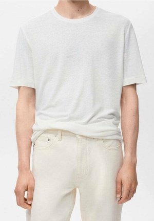 Homme portant un t-shirt blanc uni à manches courtes et un jean écru, debout avec les bras détendus le long du corps, sur un fond neutre.