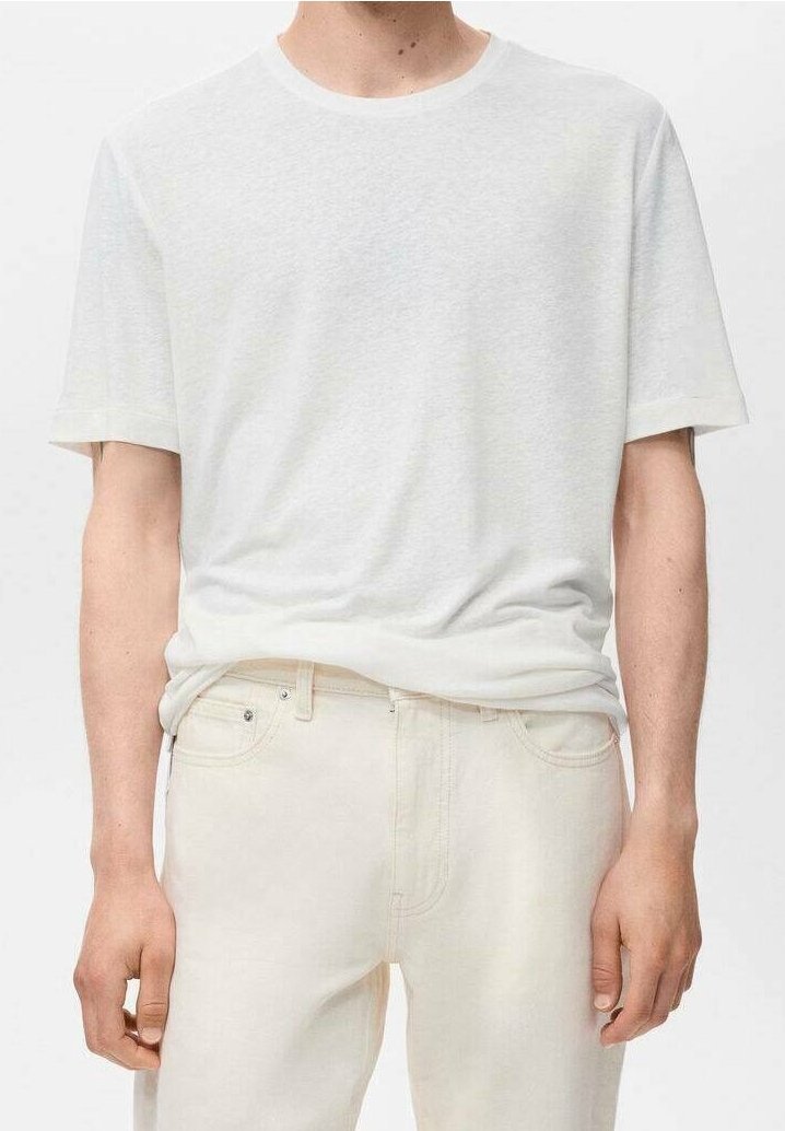 Homme portant un t-shirt blanc uni à manches courtes et un jean écru, debout avec les bras détendus le long du corps, sur un fond neutre.