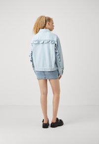 Chaqueta de mezclilla de color azul claro con ribete con volantes en los hombros y las mangas, combinada con una falda corta de mezclilla y zapatos de plataforma negros.