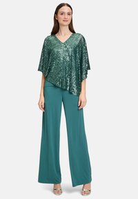 Green sequined poncho-style top with a V-neck, paired with loose, teal wide-leg trousers and silver open-toe shoes.