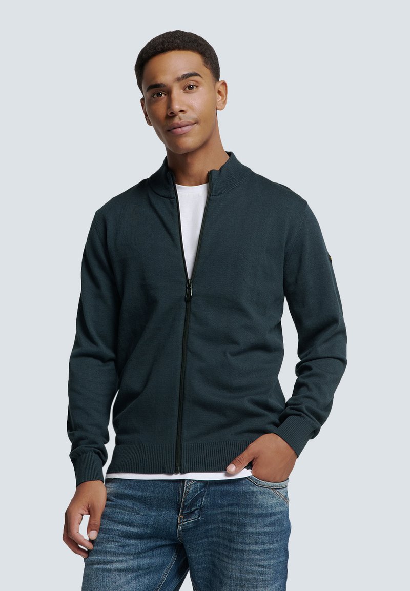 Donkergroene ritsvest met een hoge kraag, geribbelde manchetten en zoom, gemaakt van gebreide stof. Draagt over een wit T-shirt met blauwe jeans.