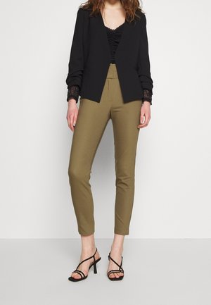 Femme portant un pantalon ajusté vert olive, un haut en dentelle noir, un blazer noir et des sandales à talons hauts noires à brides, debout sur un fond uni.