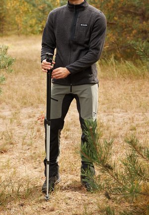 Person trägt Outdoor-Wanderausrüstung, hält zwei schwarze Trekkingstöcke und steht auf einem trockenen, grasbewachsenen Pfad umgeben von Kiefern.