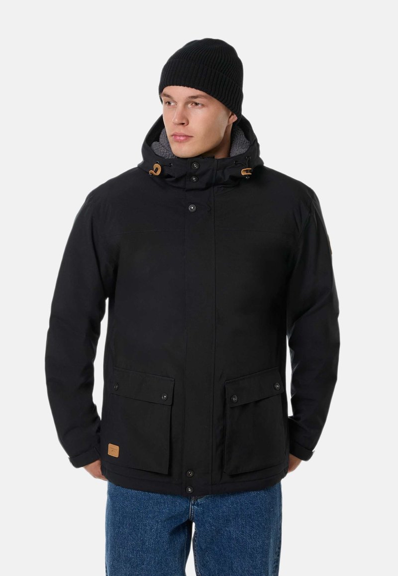 Uomo con giacca invernale nera con tasche frontali e cappuccio, berretto di lana nero e jeans blu, che guarda di lato.