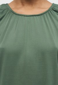 Gros plan d'une personne portant un haut vert à épaules dénudées avec des fronces le long de l'encolure.