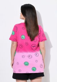 Camiseta rosa con patrones de caras sonrientes contorneadas en negro en rosa y verde. Manga corta, ajuste relajado y un efecto degradado en el dobladillo.