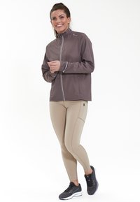 Letvægts gråbrun zip-up jakke med reflekterende accenter, kombineret med beige leggings med sidelommer og sorte atletiske sko.