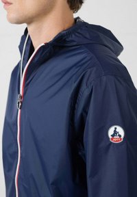 Marineblaue Kapuzenjacke aus leichtem Stoff. Verfügt über einen kontrastierenden weißen und roten Reißverschluss sowie ein rundes Patch-Logo auf dem linken Ärmel.