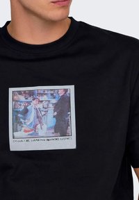 Camiseta de algodón negra con un estampado gráfico rectangular de dos figuras y el texto "¿PODRÍA LLEVAR MÁS ROPA?" debajo.