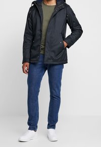 Sort vandtæt jakke med hætte, lynlås lukning, frontlommer, båret over en grøn skjorte og blå jeans, parret med hvide sneakers.