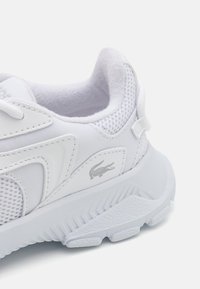 Zapatilla deportiva blanca con parte superior de malla texturizada, acentos sintéticos lisos y suela de goma con un patrón en zigzag. Detalle del logo en plata.