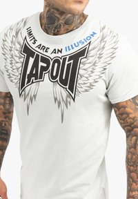 Weißes T-Shirt mit kurzen Ärmeln, das ein schwarzes "TAPOUT"-Logo mit grauen Flügeln zeigt. Der Text lautet "LIMITEN SIND EINE Illusion." Glatte Baumwolltextur.