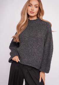 Kobieta o długich blond włosach, ubrana w ciemnoszary, teksturowany, oversize'owy sweter oraz czarne plisowane spodnie, pozująca z jedną ręką w kieszeni.