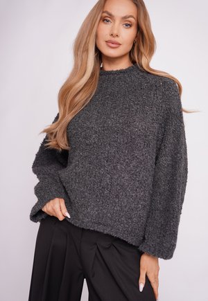 Frau mit langen blonden Haaren, die einen dunkelgrauen, strukturierten, übergroßen Pullover und schwarze, plissierte Hosen trägt und mit einer Hand in der Tasche posiert.