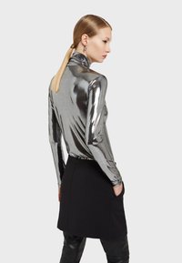 Metallisch silbernes langärmliges Oberteil mit hohem Kragen; das figurbetonte Design steht im Kontrast zu einem schwarzen Rock mit Taschen und betont die eleganten Texturen.