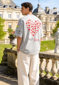 Jeune homme portant un t-shirt gris avec un motif cœur rouge et le texte « Sweet heart », debout près d'une balustrade en pierre devant un bâtiment historique.