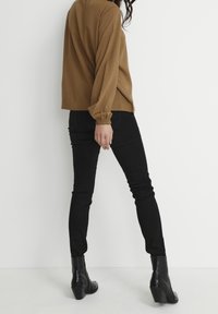 Blusa marrone a maniche lunghe con polsini elastici abbinata a jeans skinny neri e stivaletti neri con tacco a campana, vista da dietro.