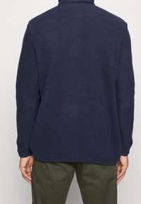 Chaqueta de forro polar azul marino con cuello alto, textura suave y corte relajado. Cuenta con mangas largas y un dobladillo redondeado.