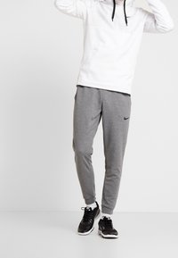Bílá mikina s kapucí a černými akcenty, šedé tepláky s logem Nike. Černé sportovní boty dokončují celý outfit.