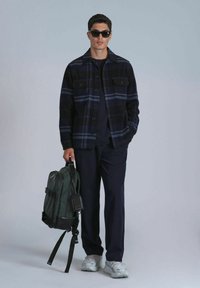 Chemise en flanelle à carreaux noirs et bleus, pantalon noir, sac à dos vert, chaussures grises. Les accessoires comprennent des lunettes de soleil noires et une étiquette noire sur le sac.
