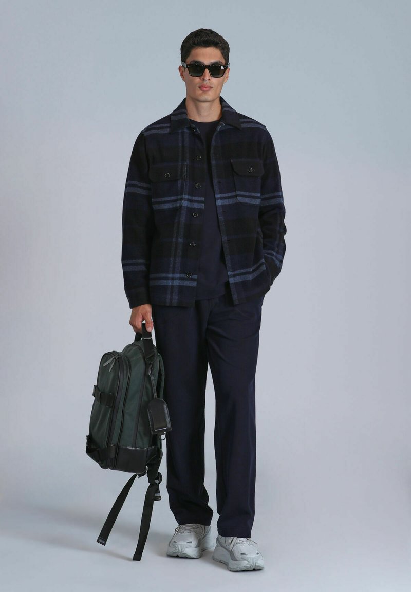 Chemise en flanelle à carreaux noirs et bleus, pantalon noir, sac à dos vert, chaussures grises. Les accessoires comprennent des lunettes de soleil noires et une étiquette noire sur le sac.