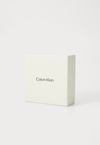 Calvin Klein kutija, kvadratnog oblika, glatka površina boje kreme, suptilan logo otisnut u smeđoj boji, minimalistički dizajn bez dodatnih akcenta.