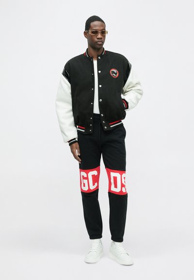 Men's black and white varsity jacket with red accents, paired with black pants featuring bold red and white lettering. White sneakers complete the outfit.