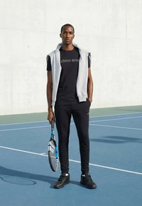 Άνδρας που στέκεται σε γήπεδο τένις με μπλε και λευκή ρακέτα τένις, φορώντας μαύρα αθλητικά ρούχα και γκρι φούτερ καλοκαιρινό στολίδι στους ώμους.